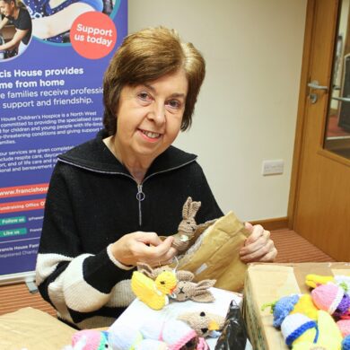 Woman wearing a black cardigan holding a knitted bunny and opening parcels of knitted items in an office