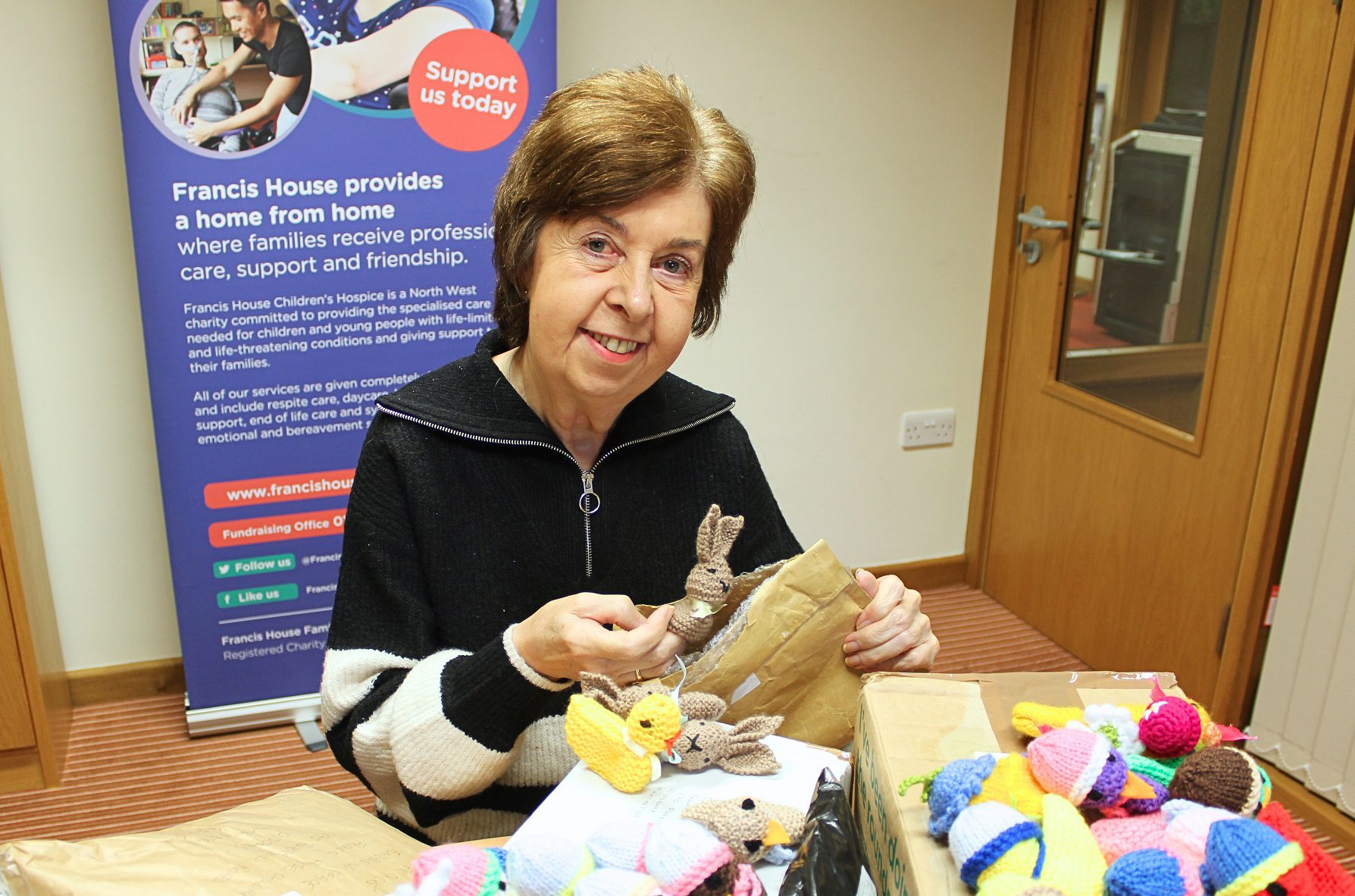 Woman wearing a black cardigan holding a knitted bunny and opening parcels of knitted items in an office