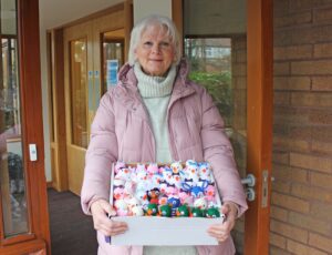 Woman wearing a pink coat and white jumper holding a box containing small knitted chicks of varying colours