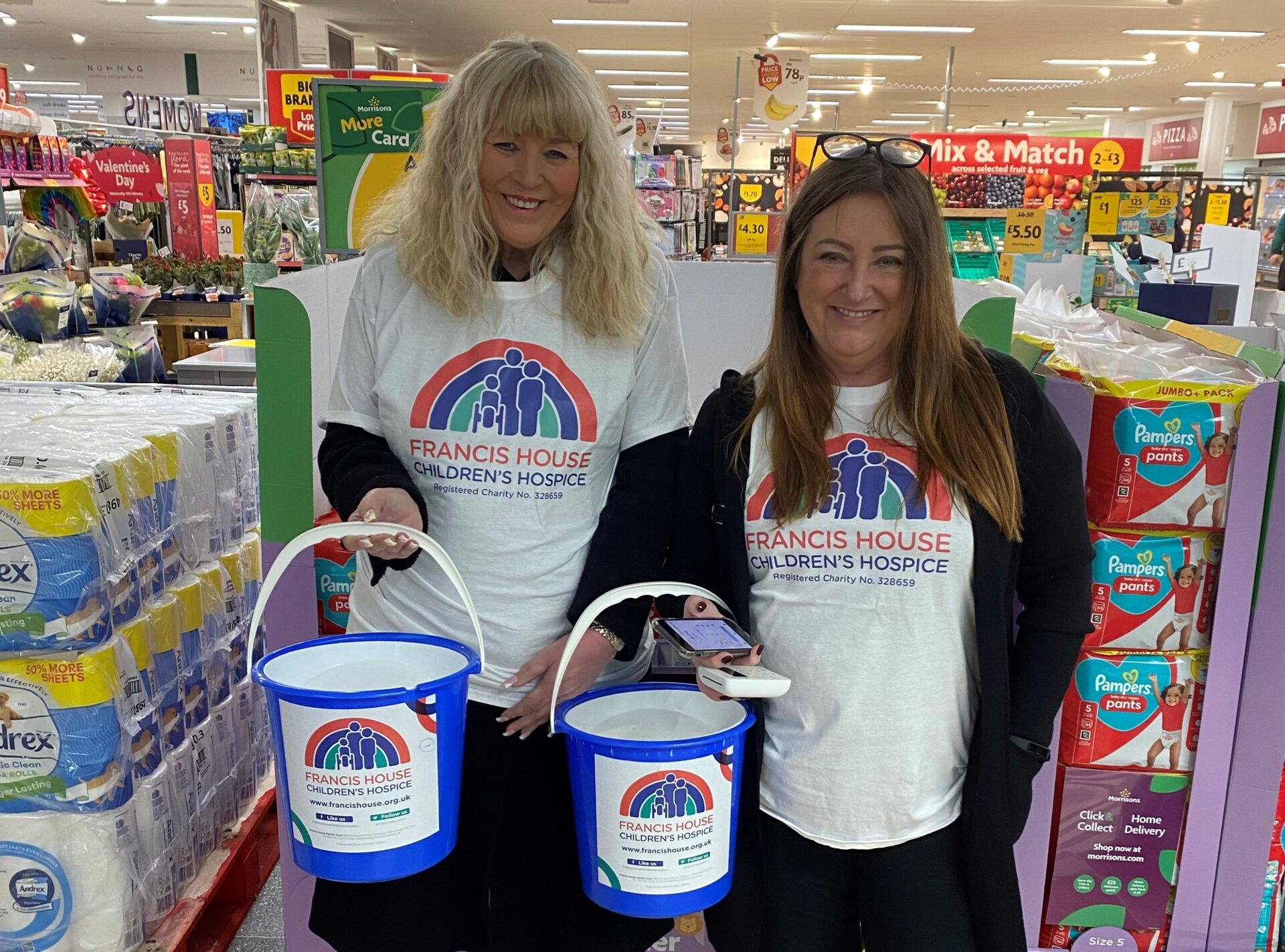 Two women wearing white t-shirts with a rainbow logo stood in a supermarket holding charity collection buckets
