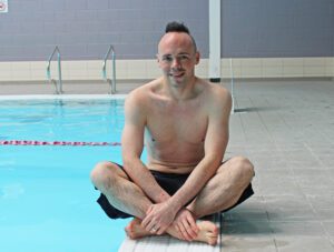 Man wearing black shorts sat cross legged at the side of swimming pool