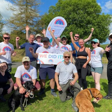 Group of men and women and pet dogs wearing white t-shirts in front of a charity flag and holding a sign saying We did it.
