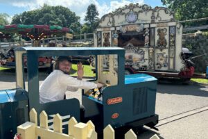 Man driving a blue train on a miniature railway with a fairground organ in the background