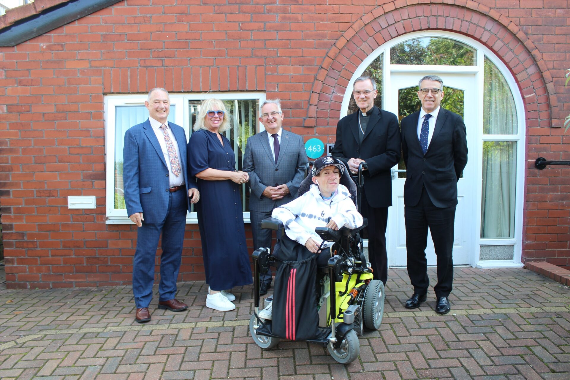 Group of men and women one in a wheelchair in front of a brick house