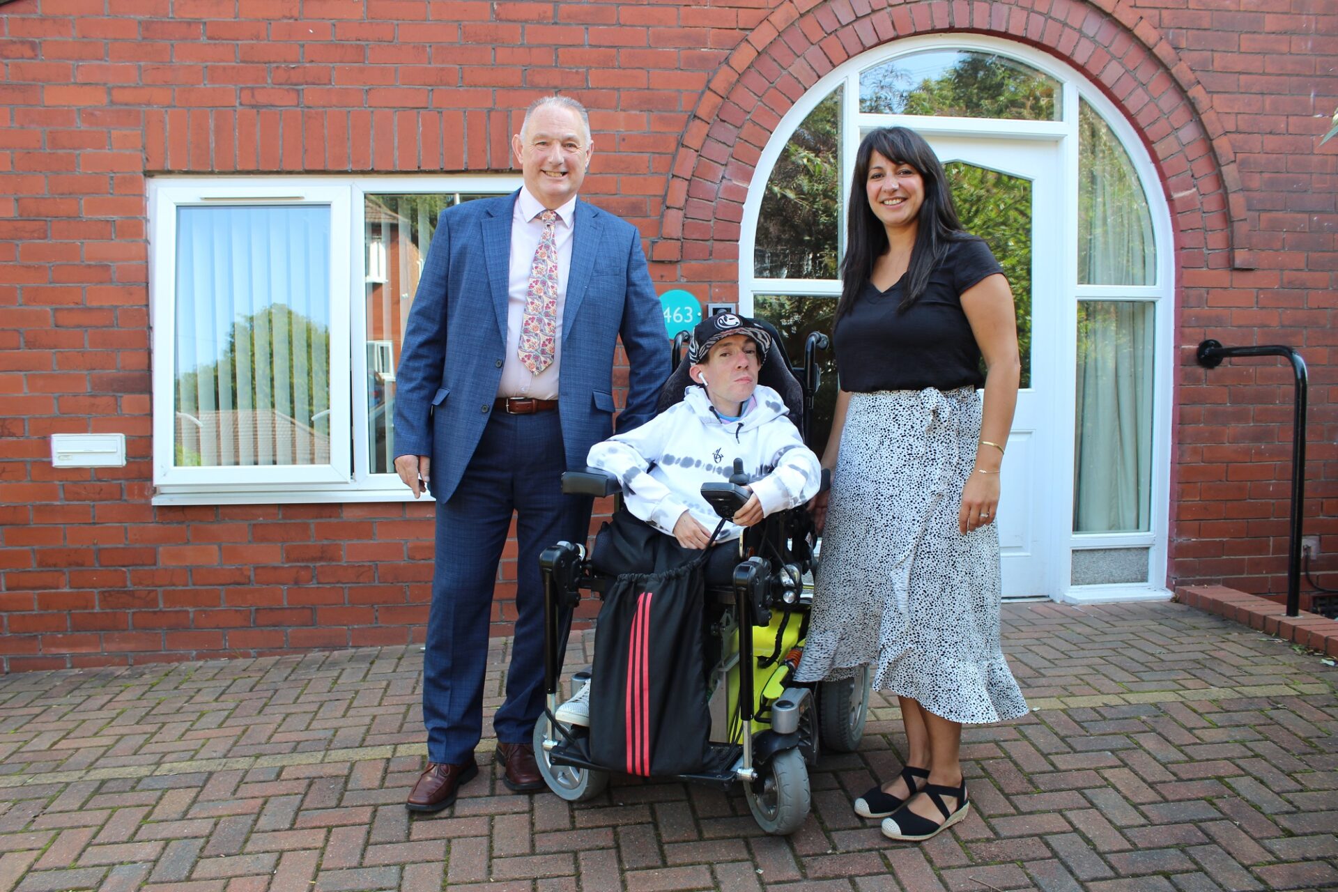 A man and a woman stood next to a young man in a wheelchair in front of a house