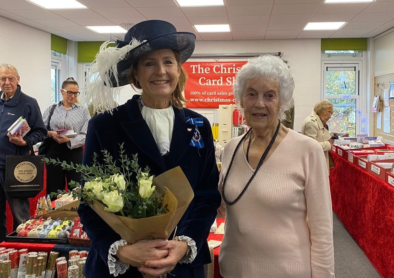Woman wearing a large hat with white feather holding a bunch of flowers stood next to a woman wearing a pink jumper in a shop