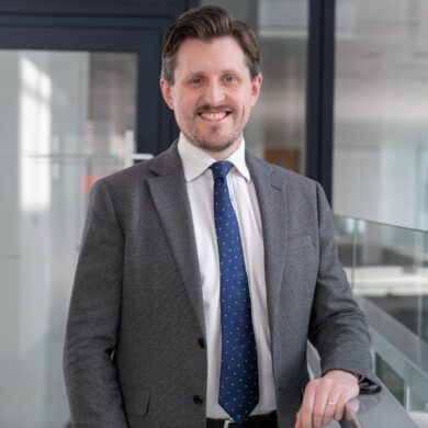 Man with a beard wearing a grey suit and blue tie stood on a glass corridor smiling at the camera