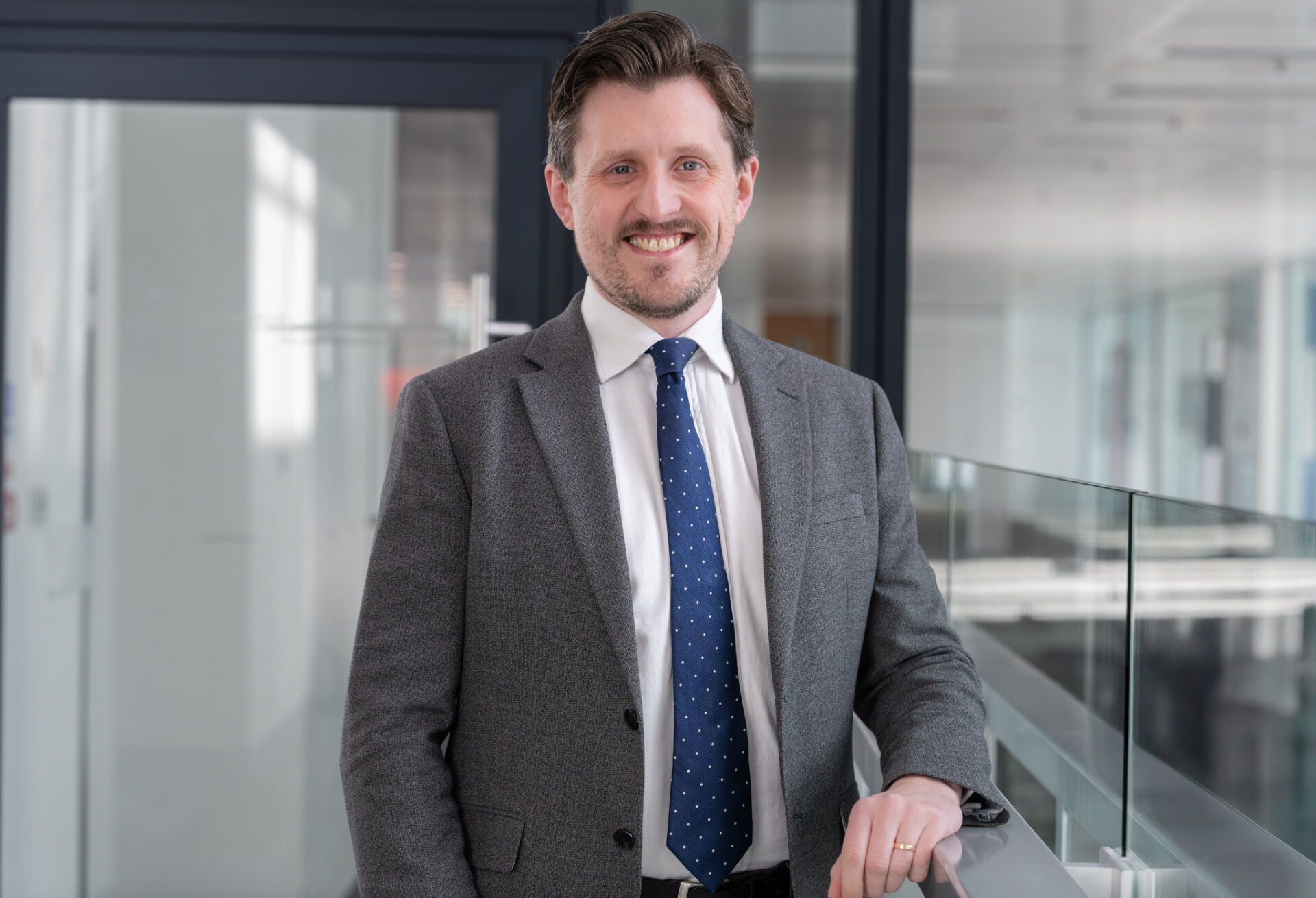 Man with a beard wearing a grey suit and blue tie stood on a glass corridor smiling at the camera