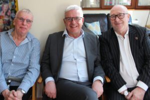 Three men seated wearing shirts and jackets smiling at the camera