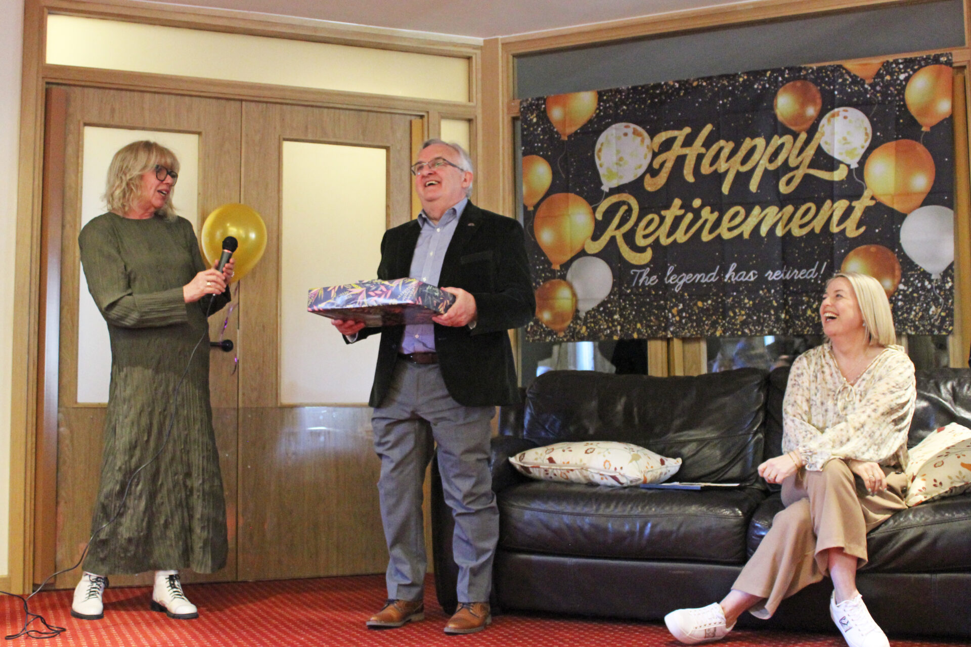 Man holding a present smiling stood in front of a banner saying happy retirement