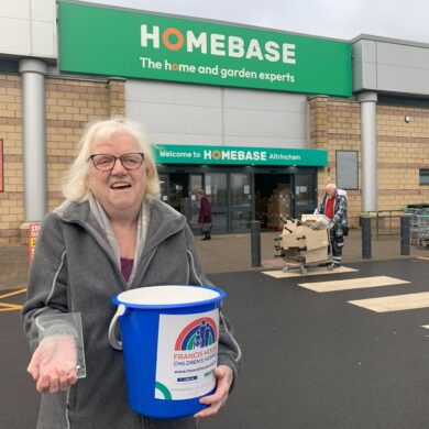 Woman with grey hair stood holding a blue charity collection bucket and a glass award trophy stood outside a large store with the words Homebase over the entrance