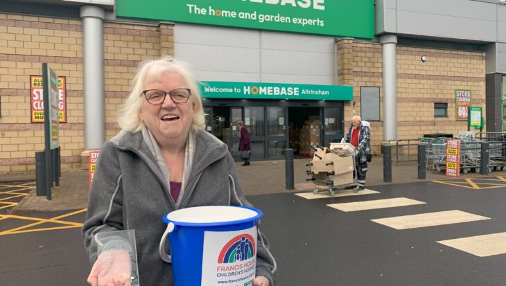 Woman with grey hair stood holding a blue charity collection bucket and a glass award trophy stood outside a large store with the words Homebase over the entrance