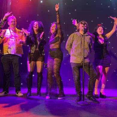 People stood on a stage dressed as various singers and artists waving their hands in the air and singing