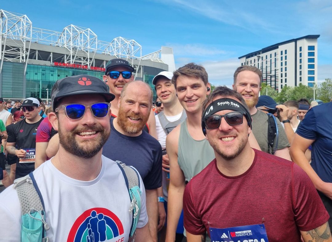 Crowd of men wearing running shirts stood smiling at the camera in front of Old Trafford football ground