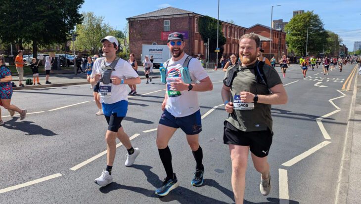 Three men running on the road with other runners around them smiling at the camera
