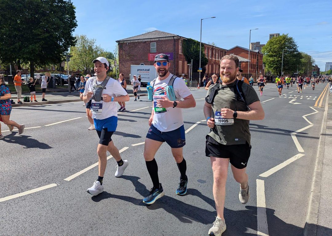 Three men running on the road with other runners around them smiling at the camera