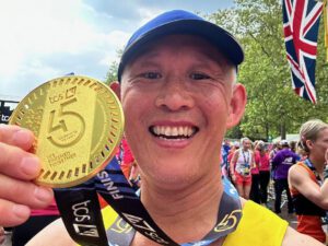 Man wearing a blue cap and yellow vest smiling at the camera holding a gold London Marathon medal