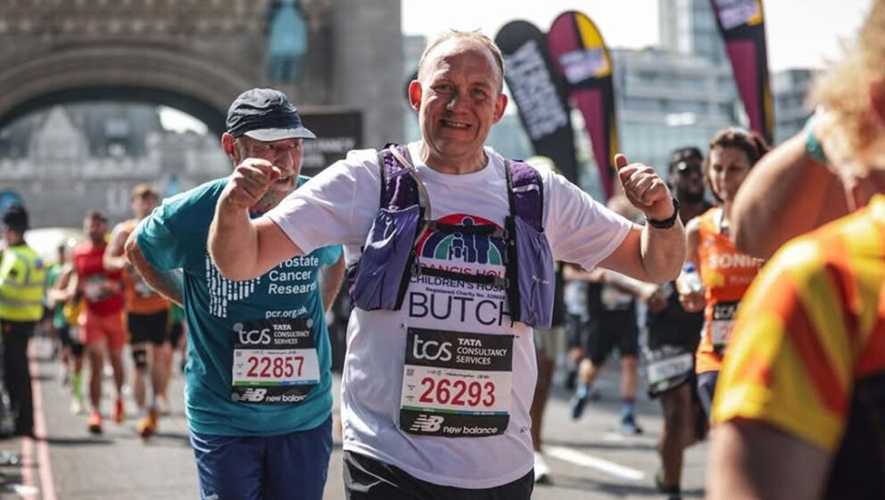Man wearing a white running t-shirt holding his arms in the air crossing Tower Bridge with other runners in the London Marathon