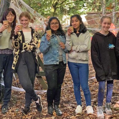 Group of young people stood in a forest holding paper coffee cups and smiling at the camera