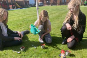 Three children sat on a lawn one of them watering small flower pots with a watering can