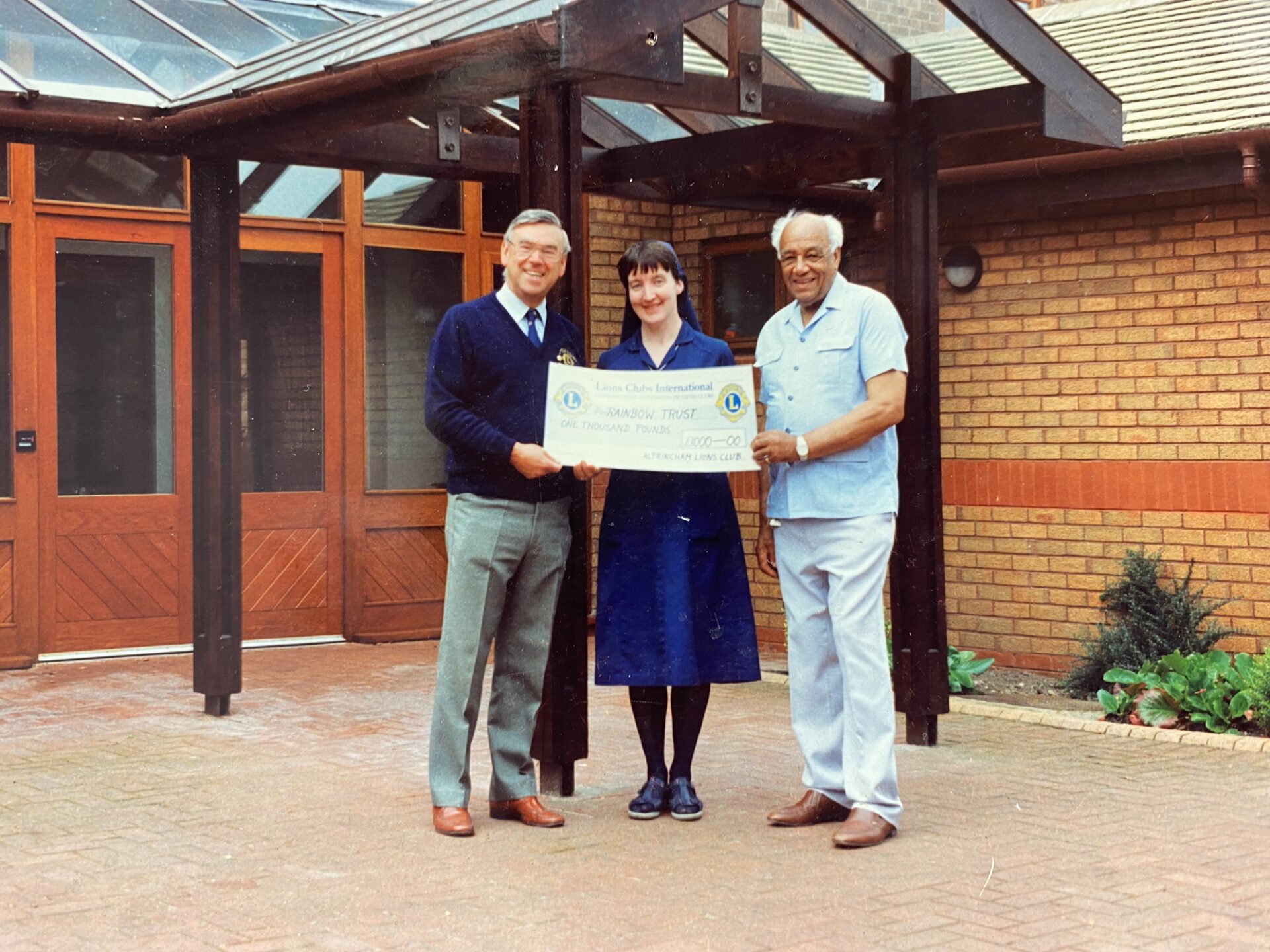 A religious sister being handed a large cheque by two men stood outside a building that is to become a children's hospice