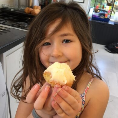 Young girl with brown shoulder length hair stood in a kitchen holding a cup cake with cream icing smiling at the camera