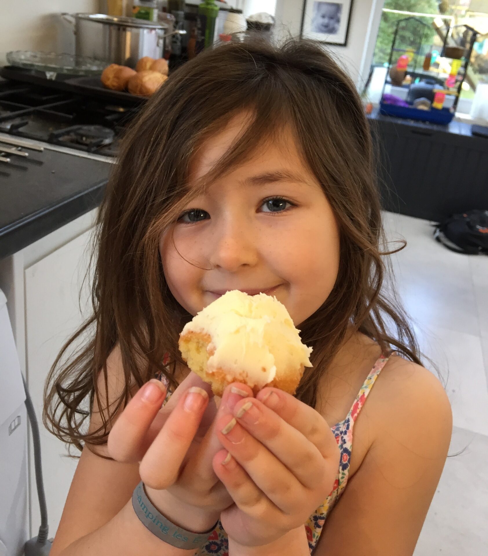 Young girl with brown shoulder length hair stood in a kitchen holding a cup cake with cream icing smiling at the camera