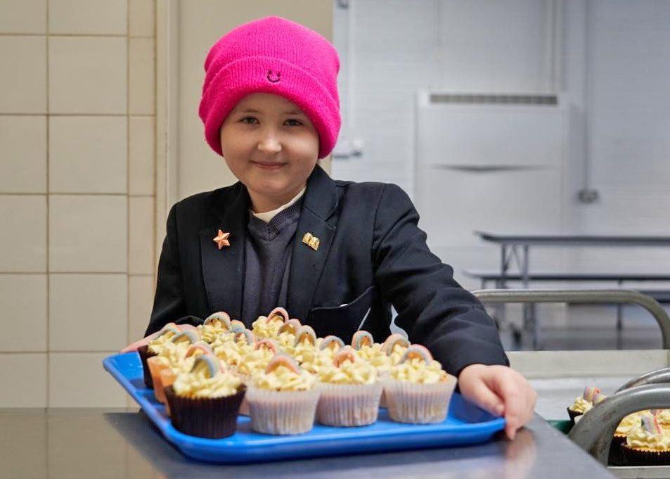 Young girl wearing a school uniform and a pink beanie hat stood in a school canteen holding a tray of cup cakes and smiling at the camera