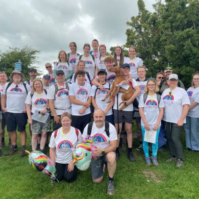 large group of people wearing walking boots and matching white charity t-shirts stood on a grassy hill smiling at the camera