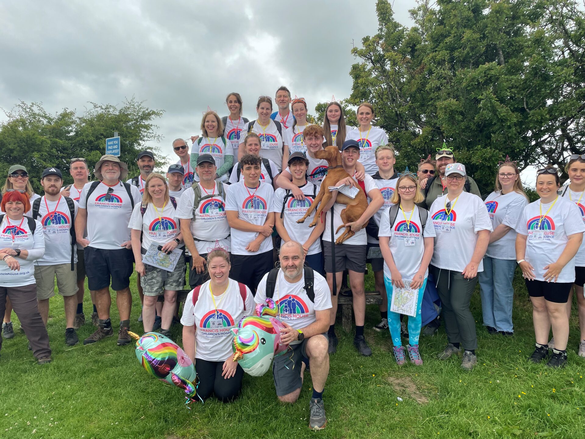 large group of people wearing walking boots and matching white charity t-shirts stood on a grassy hill smiling at the camera