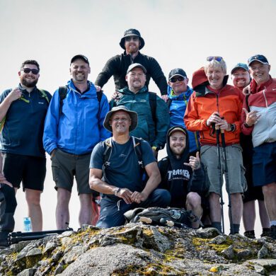 Group of men wearing hiking clothing stood at the top of a mountain smiling at the camera
