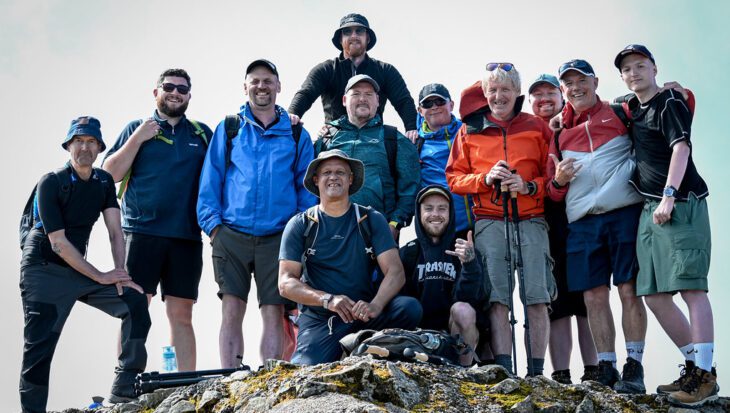 Group of men wearing hiking clothing stood at the top of a mountain smiling at the camera