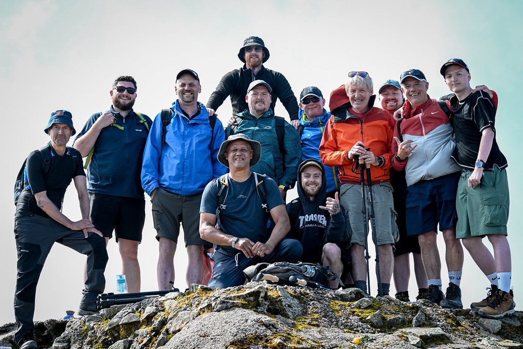 Group of men wearing hiking clothing stood at the top of a mountain smiling at the camera