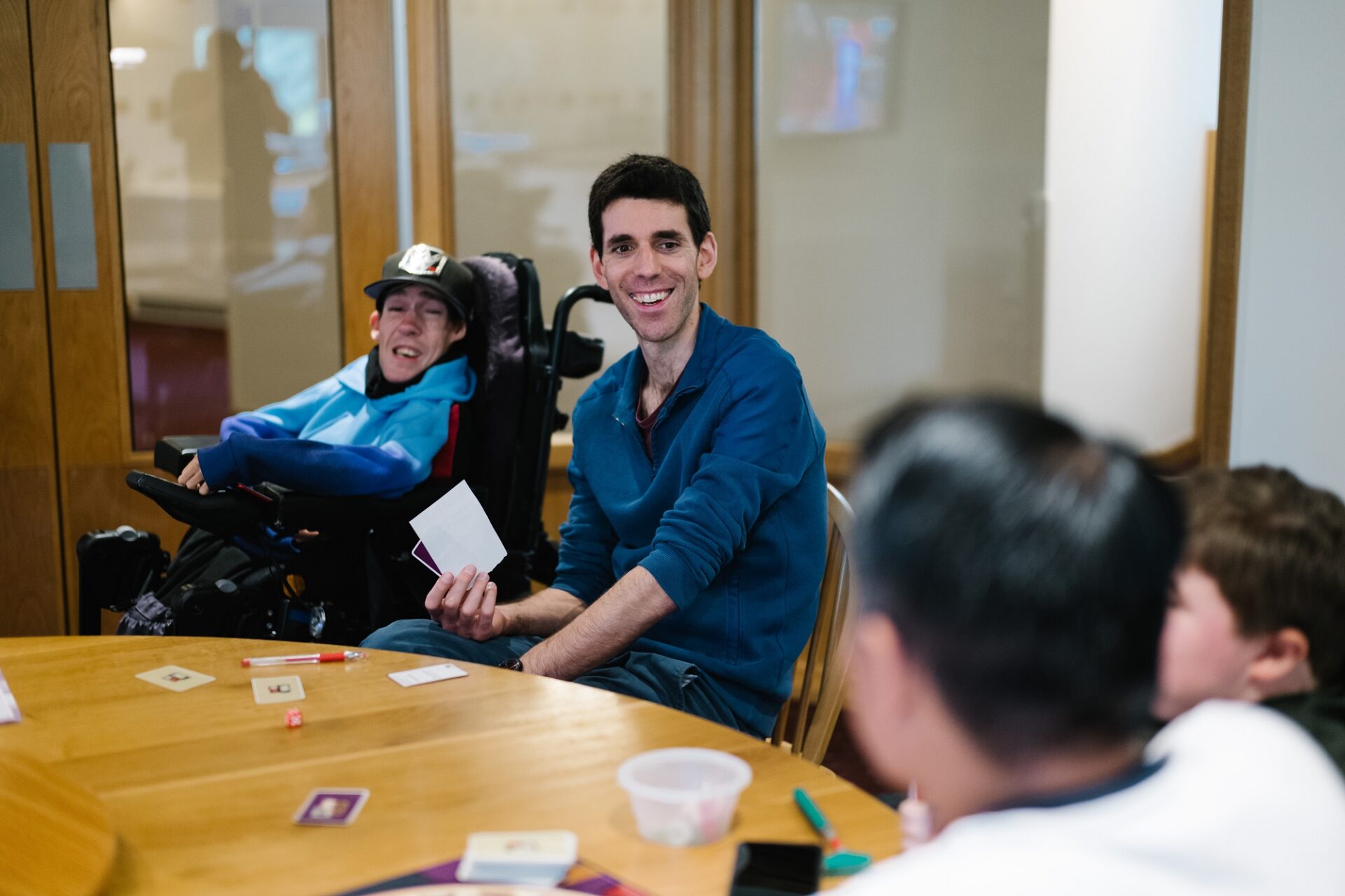 Young men playing a card game sat around a round table. One wearing a cap and sat in a wheelchair.