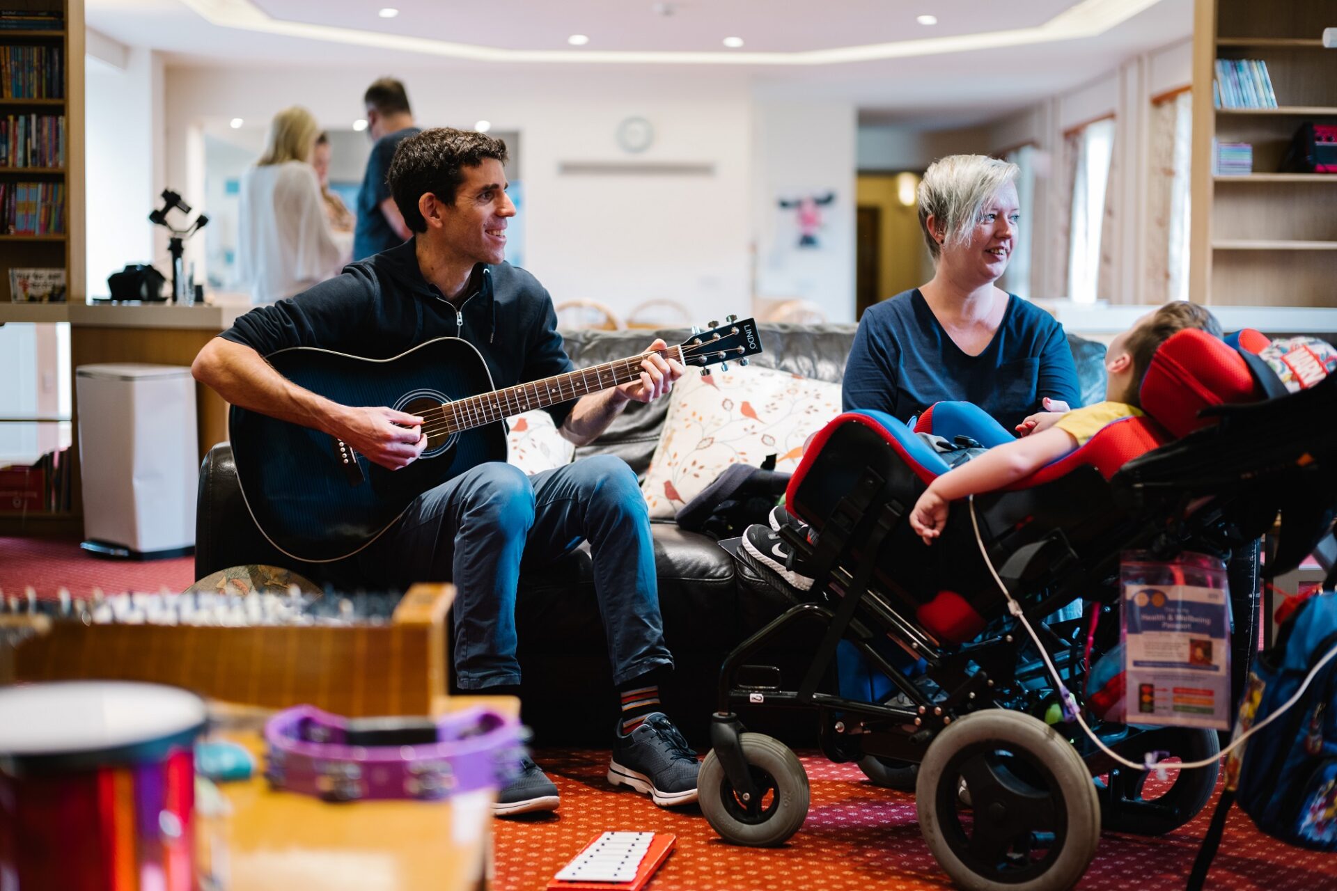Man plays a guitar sat on a sofa to a woman and young child in a wheelchair