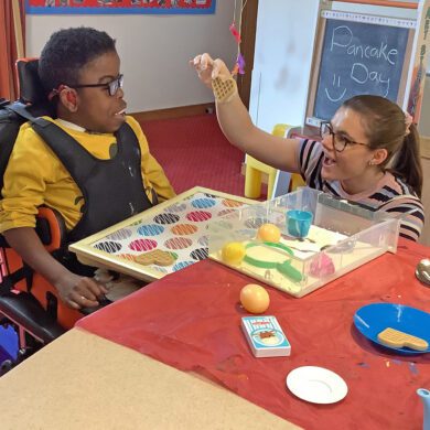Woman holding a waffle toy and showing it to a young boy sat in a wheelchair smiling