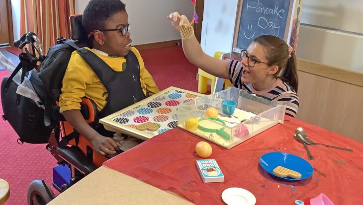 Woman holding a waffle toy and showing it to a young boy sat in a wheelchair smiling