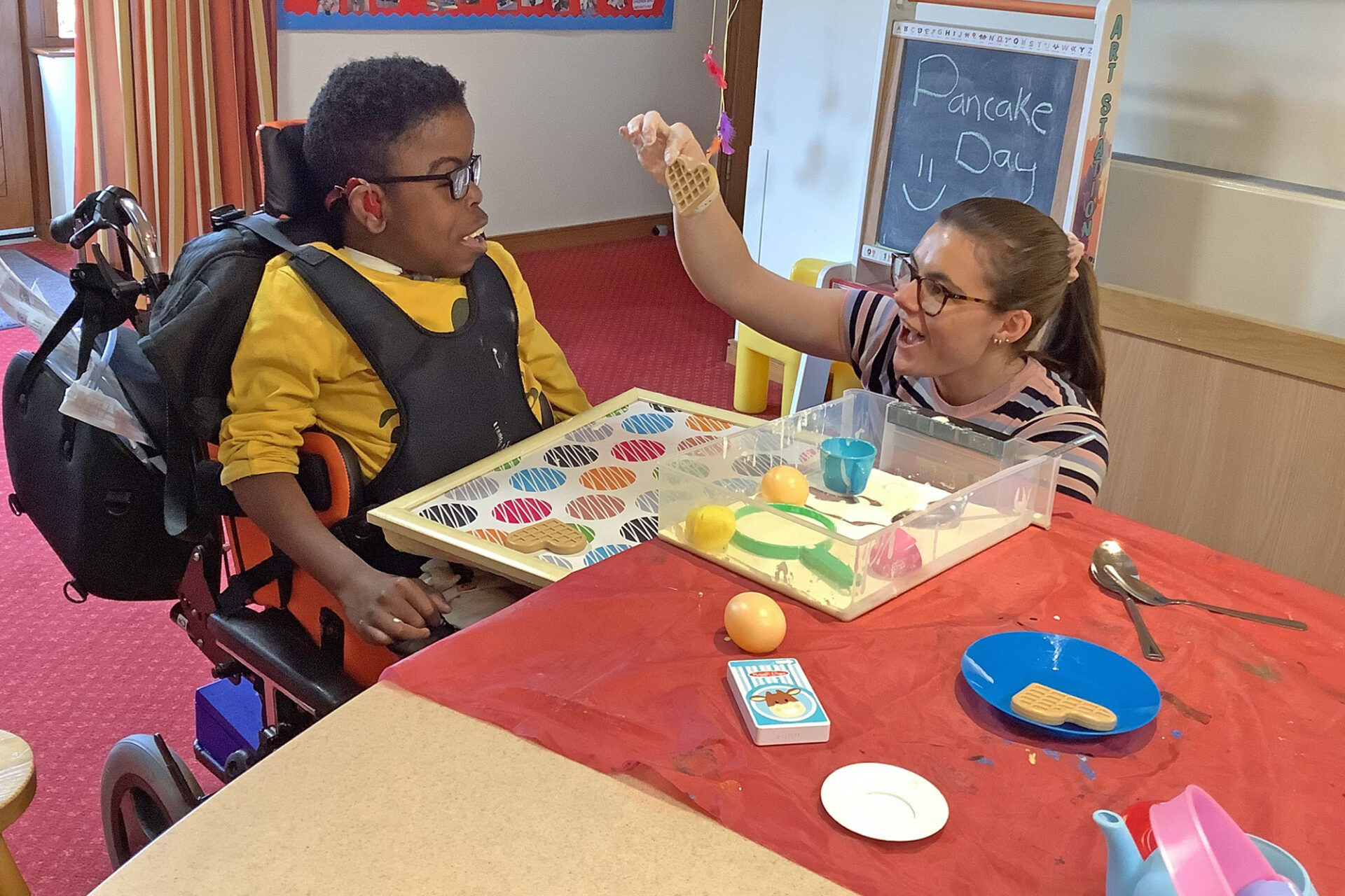 Woman holding a waffle toy and showing it to a young boy sat in a wheelchair smiling