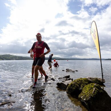A participant emerges running from the lake in wetsuit, trainers, swim cap and goggles