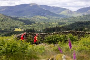 Two participants run beside a stone wall amidst greenery atop a hill with beautiful views of the countryside