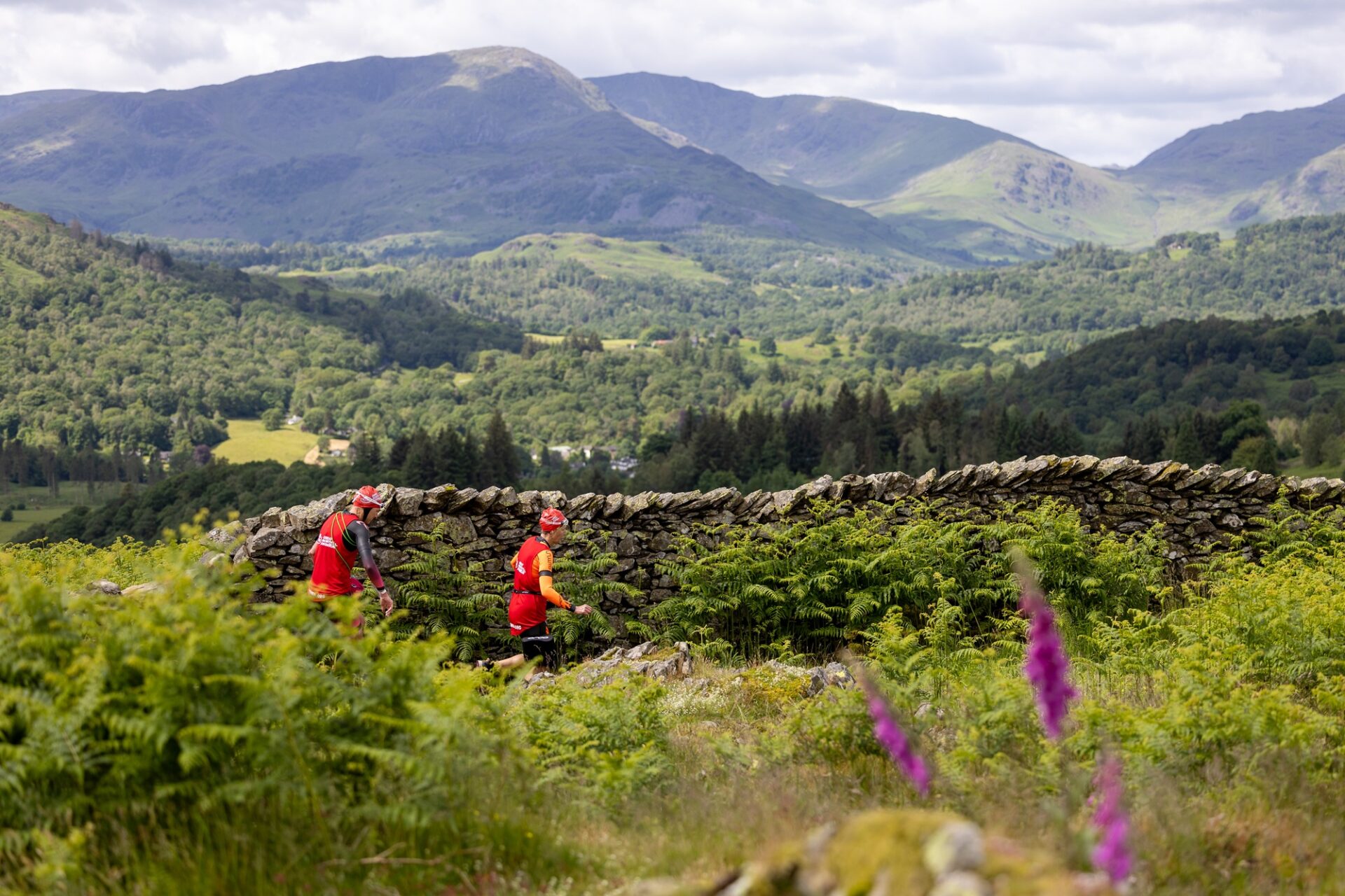 Two participants run beside a stone wall amidst greenery atop a hill with beautiful views of the countryside