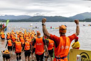 A throng of participants in Great North Swim Run tabards and wetsuits make their way into the lake to begin swimming. The nearmost participant has his arms raised in a symbol of celebration and strength! Kayaks and buoys can be seen in the water.