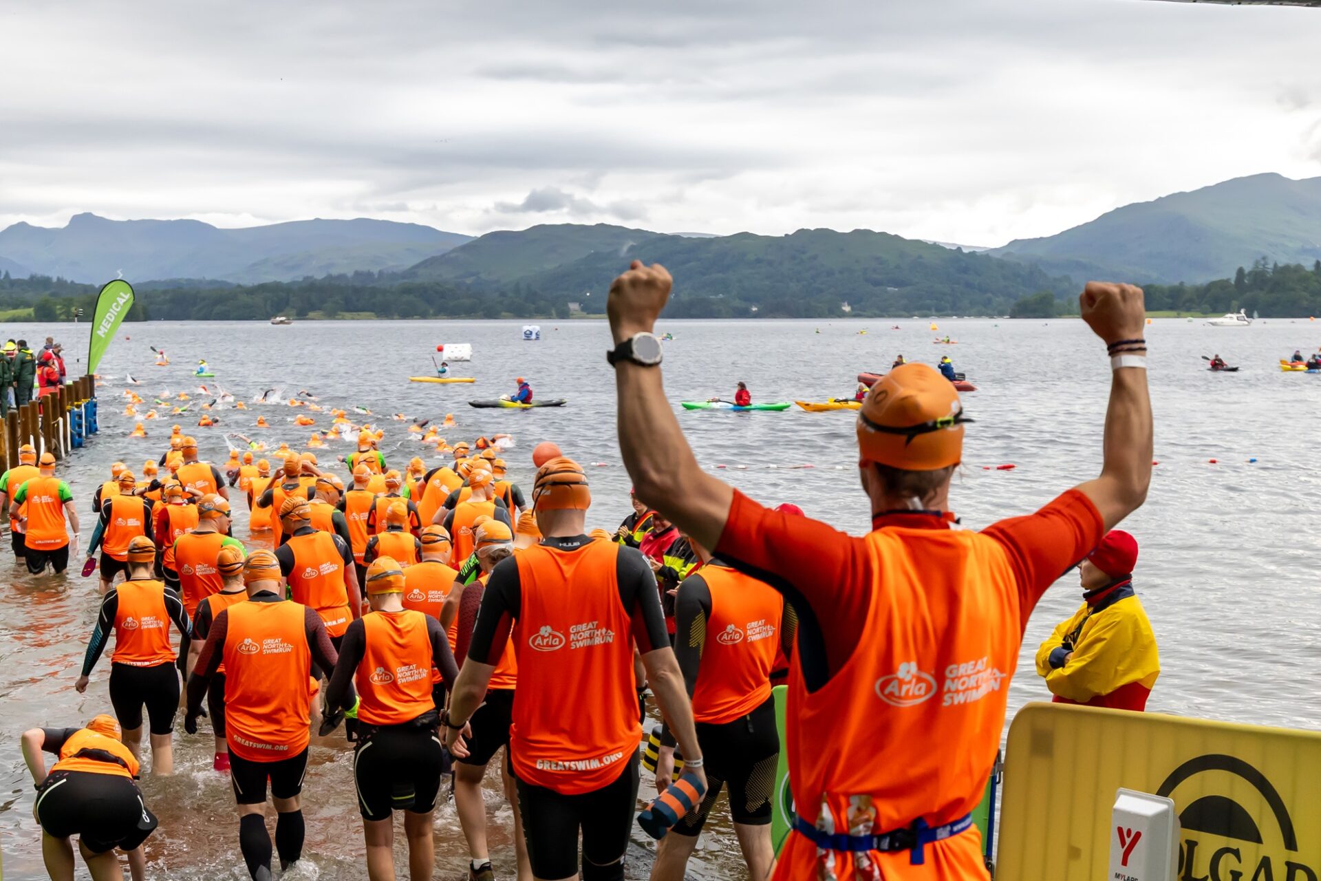 A throng of participants in Great North Swim Run tabards and wetsuits make their way into the lake to begin swimming. The nearmost participant has his arms raised in a symbol of celebration and strength! Kayaks and buoys can be seen in the water.