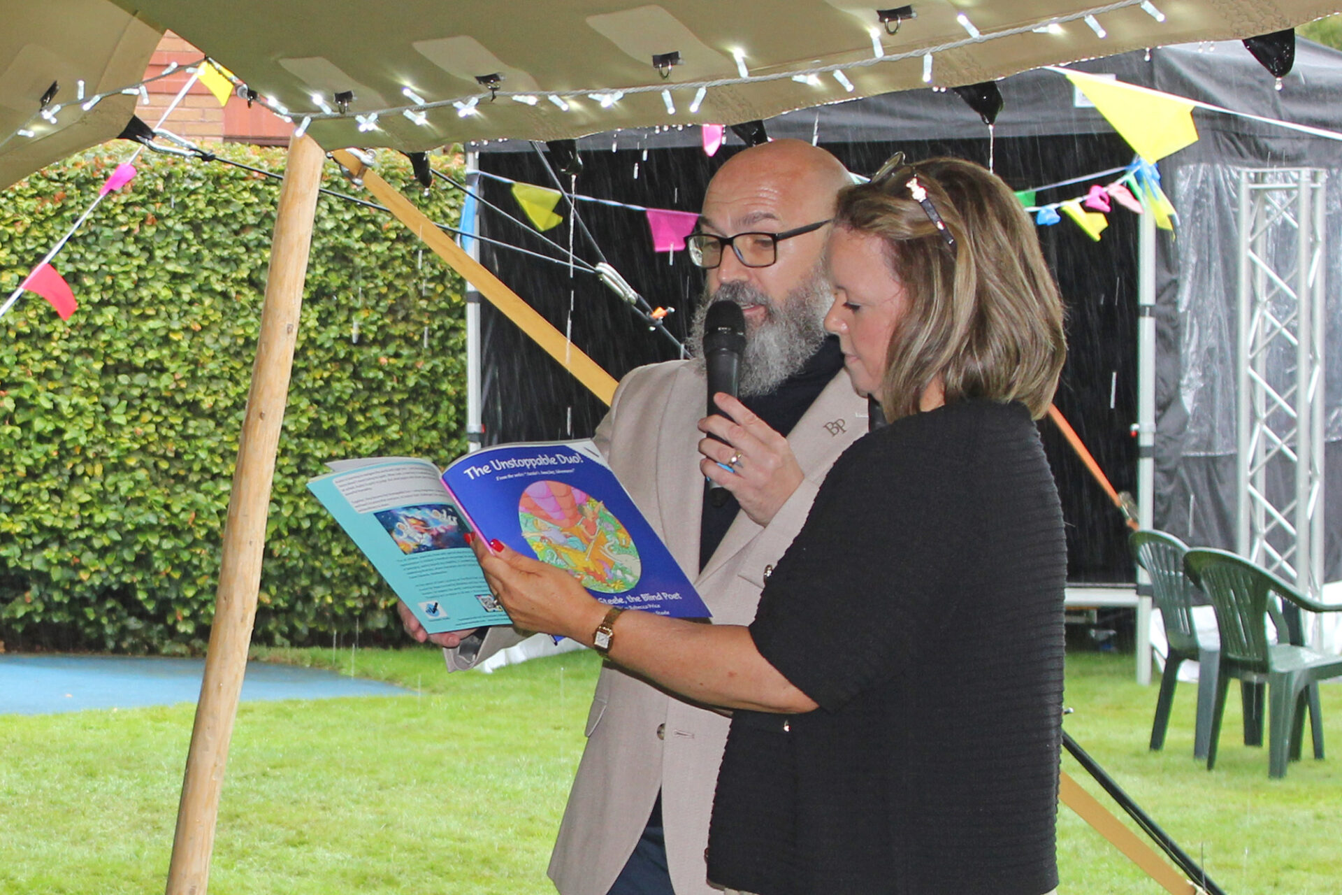 Dave Steele The Blind Poet standing alongside his wife Amy who is holding open a book The Unstoppable Duo which is being read by Dave. They stand under a tent with lights and bunting on a grassy lawn.