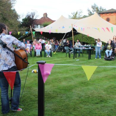 Man plays a guitar to a small audience of people sat on the lawn in front of a large tent