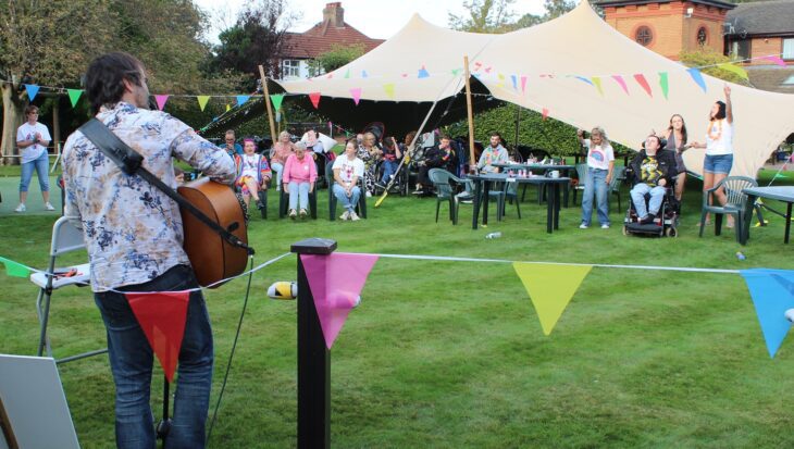Man plays a guitar to a small audience of people sat on the lawn in front of a large tent