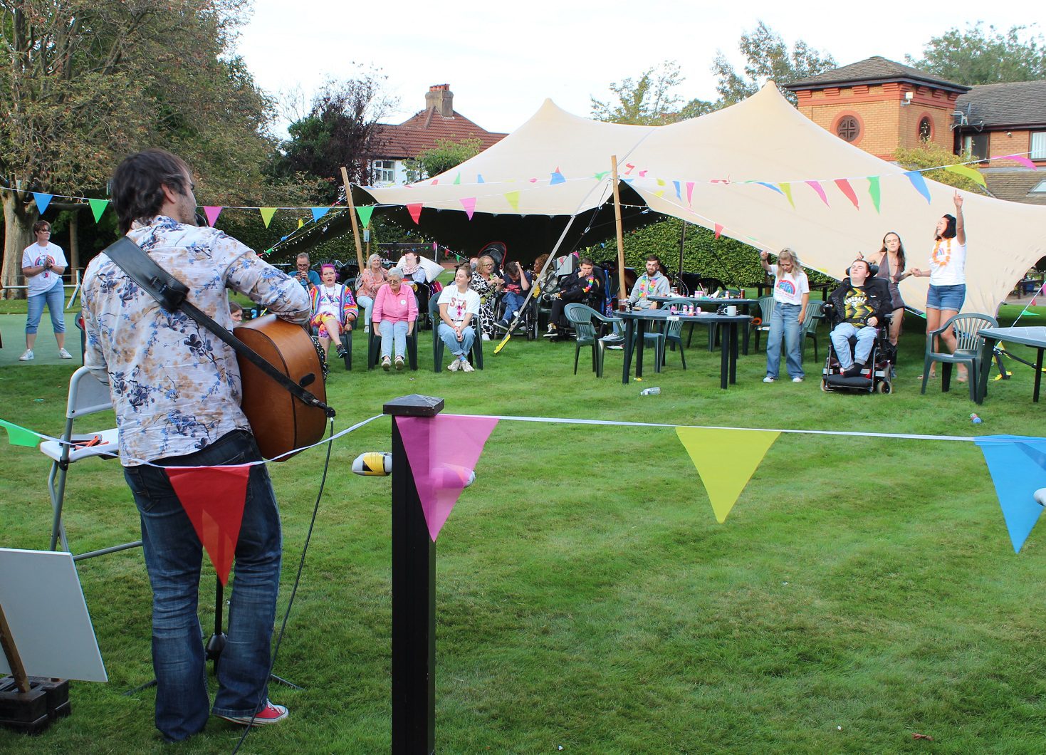 Man plays a guitar to a small audience of people sat on the lawn in front of a large tent