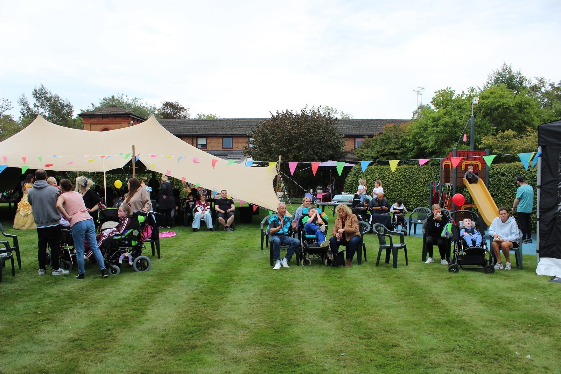 Children and families smiling and laughing sat in chairs and wheelchairs on a grassy lawn in front of a big tent decorated with colourful bunting next to a pirate ship slide.