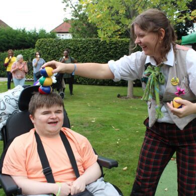 Young person in a wheelchair smiling as a woman dressed in fairground clothing balances juggling bean bags on his head. They smile at each other.