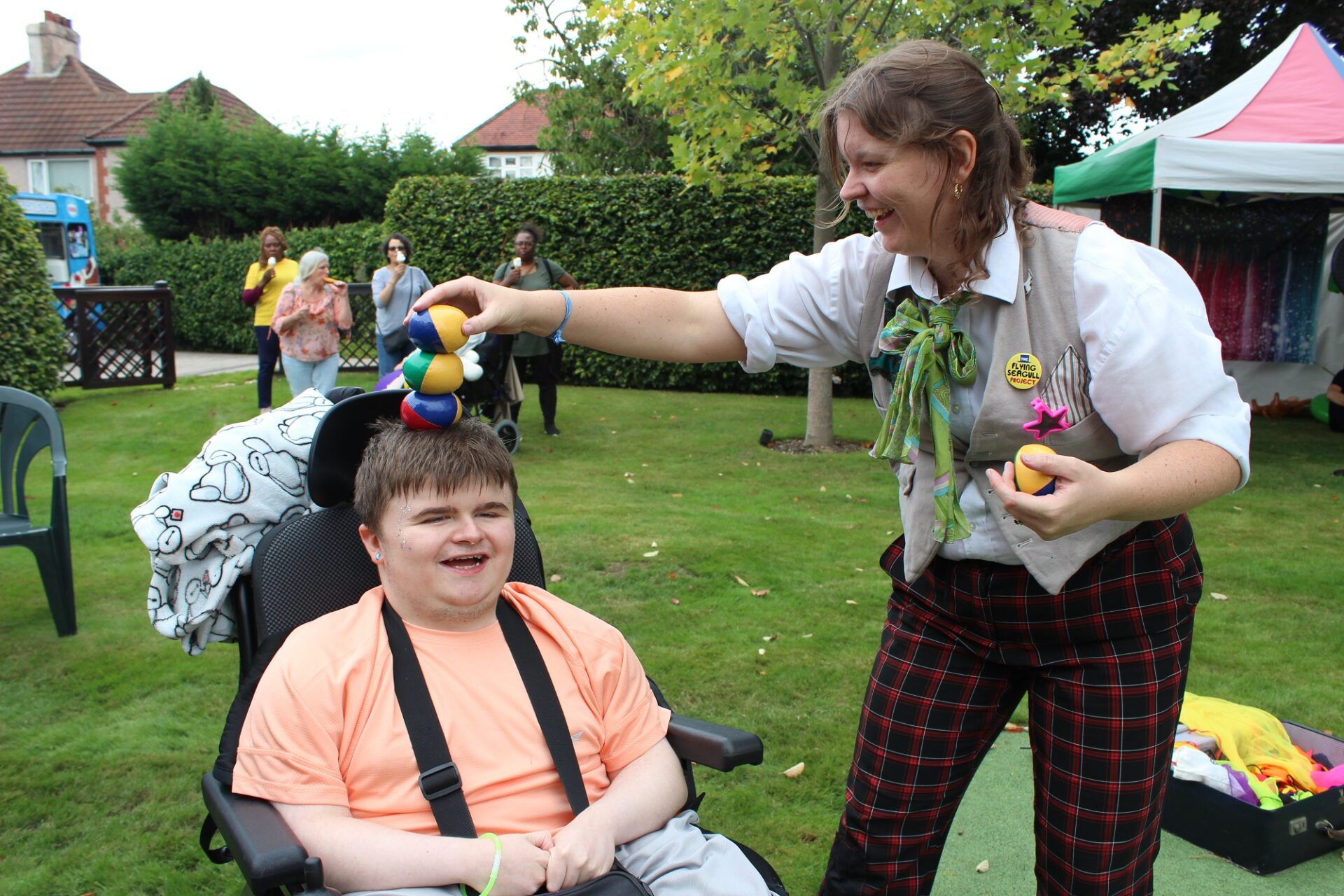 Young person in a wheelchair smiling as a woman dressed in fairground clothing balances juggling bean bags on his head. They smile at each other.